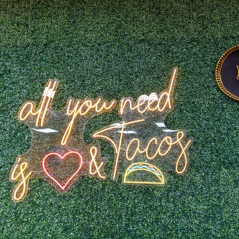 Neon sign on a green hedge wall reads "All you need is love & Tacos" with a sombrero.