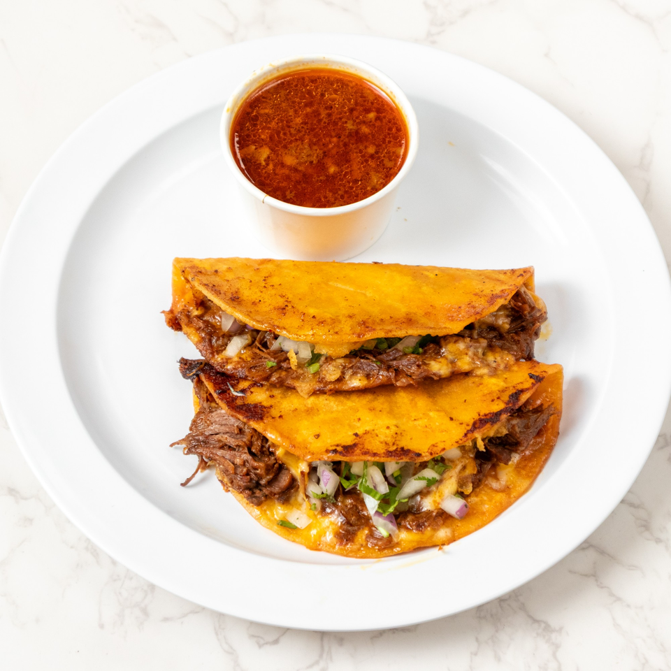 Two birria tacos with shredded beef, onions, cilantro, and melted cheese, served with a side of consommé.