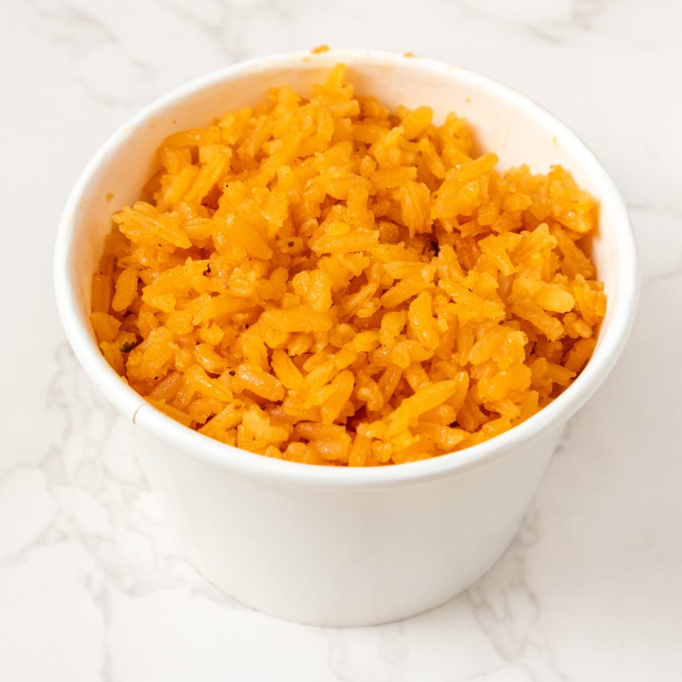 A white paper cup filled with bright orange Mexican rice on a white marble surface.