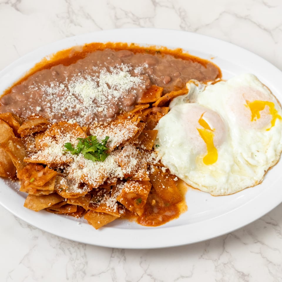 A white plate with chilaquiles, refried beans, and two fried eggs on a marble surface.