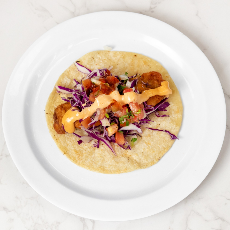 A single fish taco on a white plate, topped with red cabbage, salsa, and a creamy orange sauce.