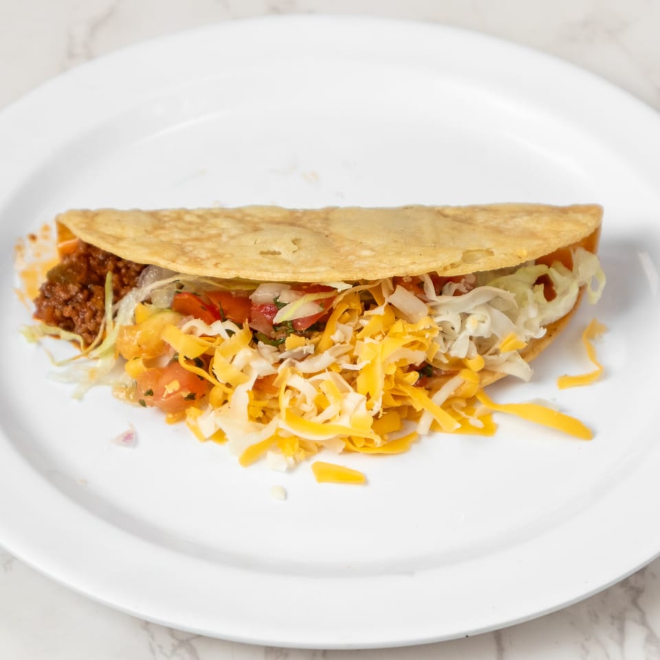 A single taco filled with ground beef, shredded cheese, lettuce, and diced tomatoes on a white plate.