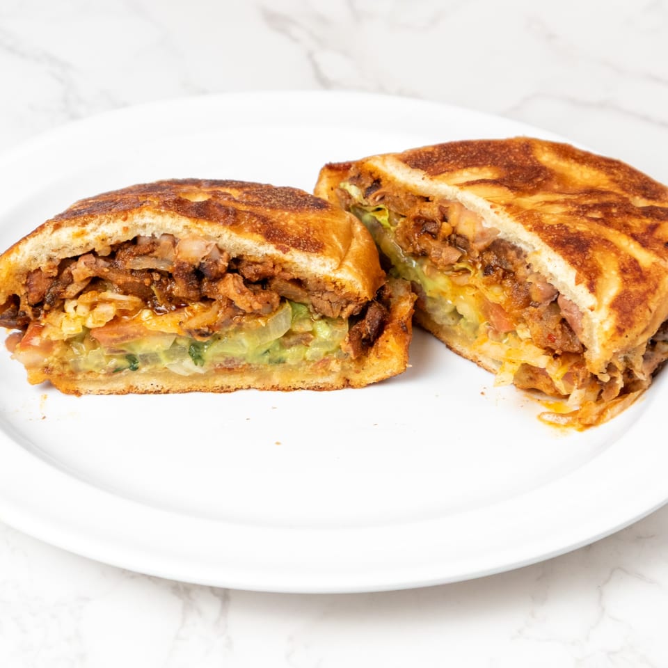 A grilled sandwich, cut in half, revealing layers of meat, guacamole, and vegetables on a white plate.