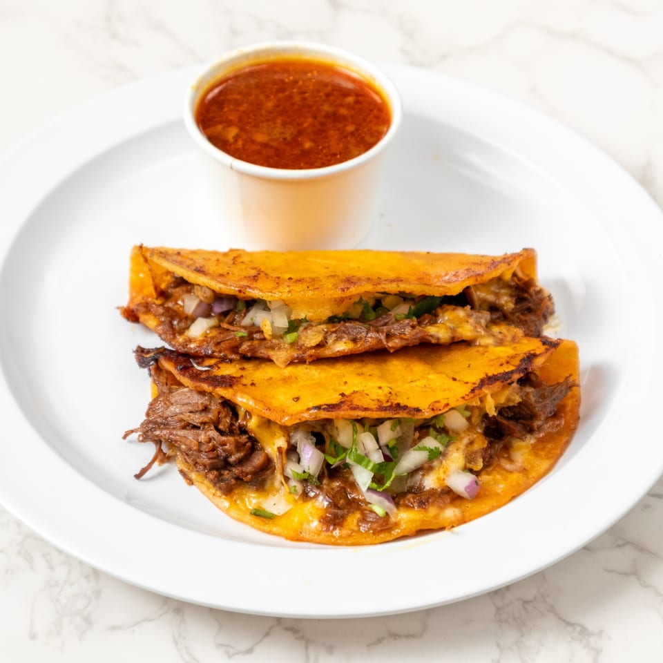 Two birria tacos with shredded beef, onions, and cilantro, served with a side of consommé.