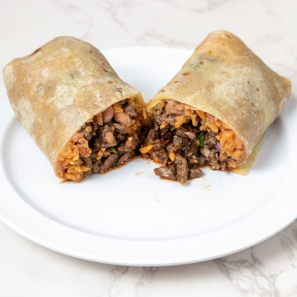 A large burrito, cut in half, revealing a filling of seasoned meat, rice, and beans on a white plate.