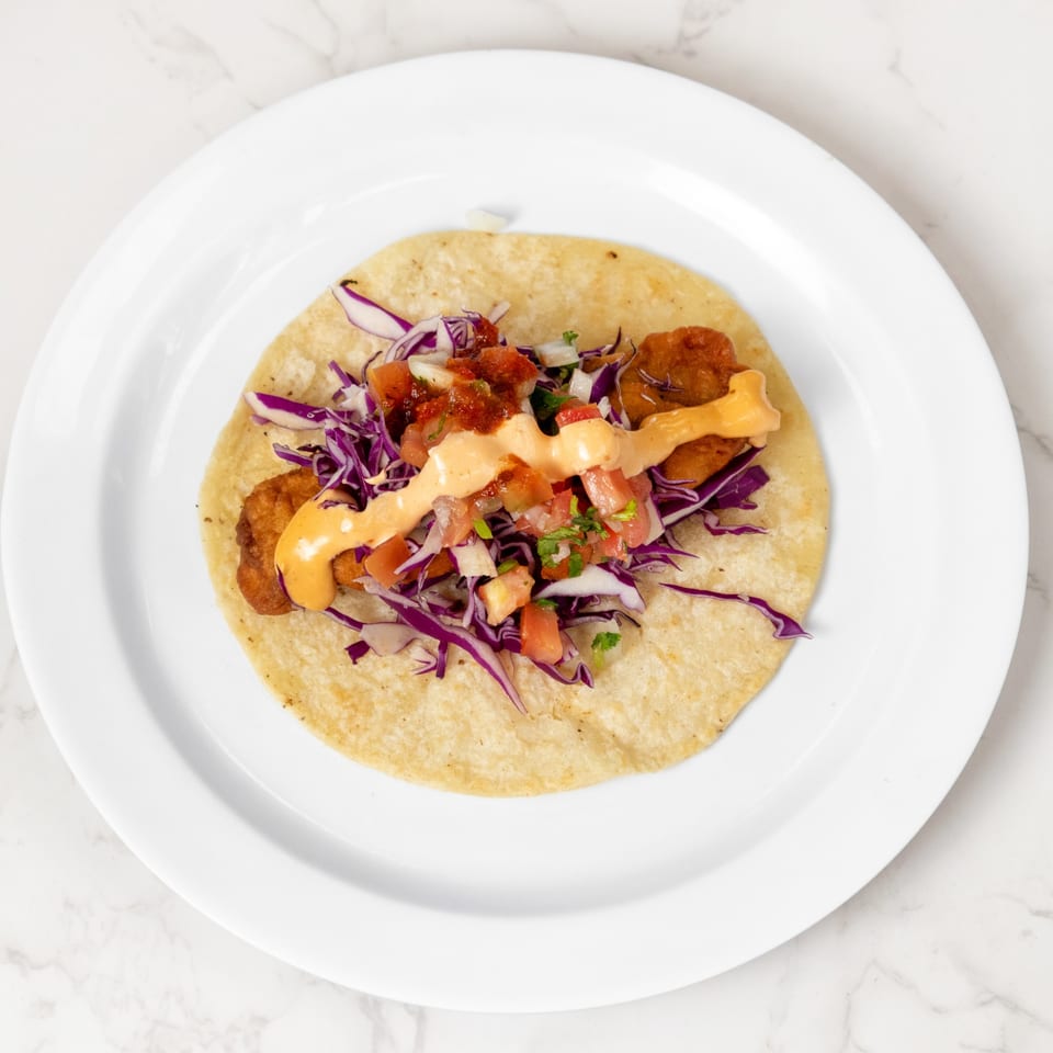 A single fish taco with red cabbage, pico de gallo, and a creamy orange sauce on a white plate.