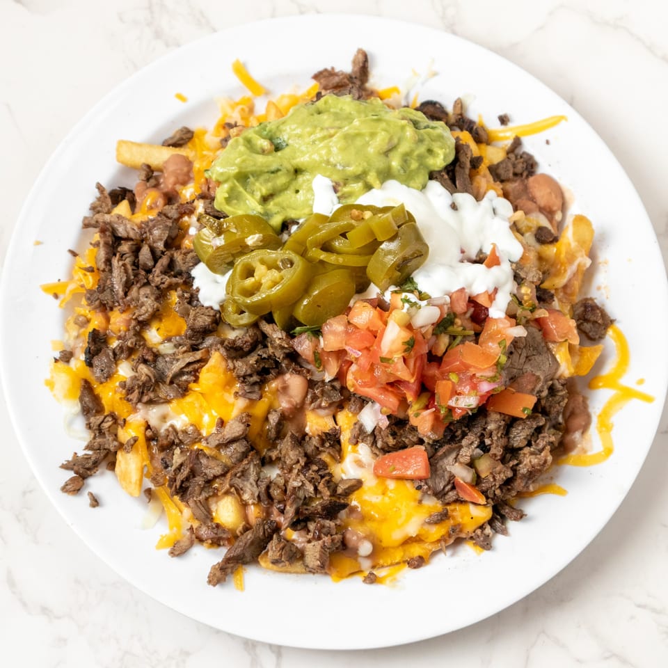 A white plate piled high with carne asada fries, topped with guacamole, sour cream, jalapeños, and pico de gallo.