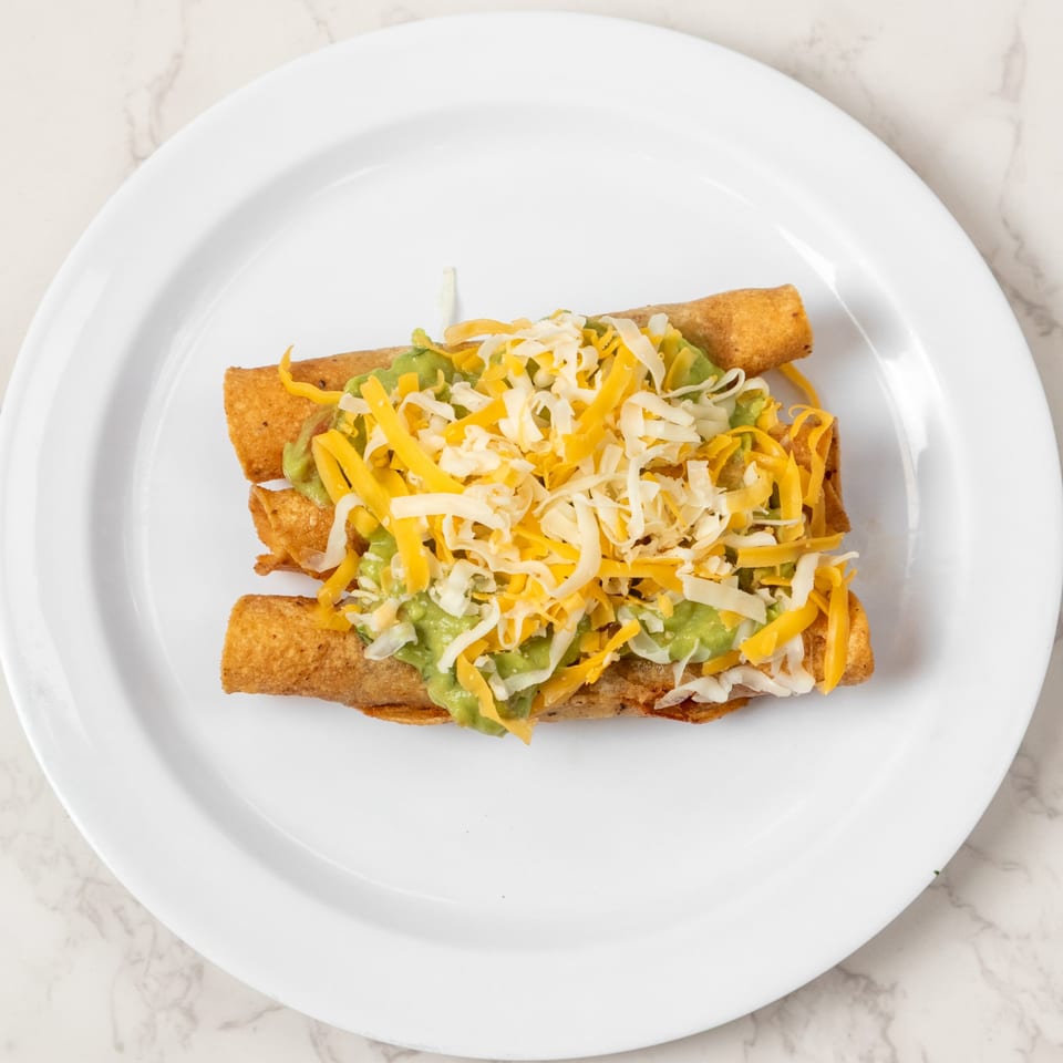 Three golden taquitos topped with guacamole, shredded cheddar, and Monterey Jack cheese on a white plate.