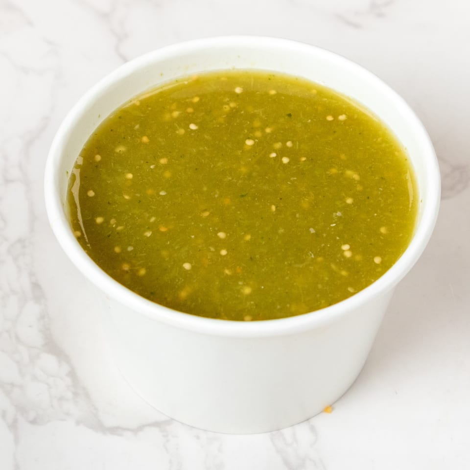 A white cup filled with vibrant green salsa verde, speckled with white seeds, on a marble surface.