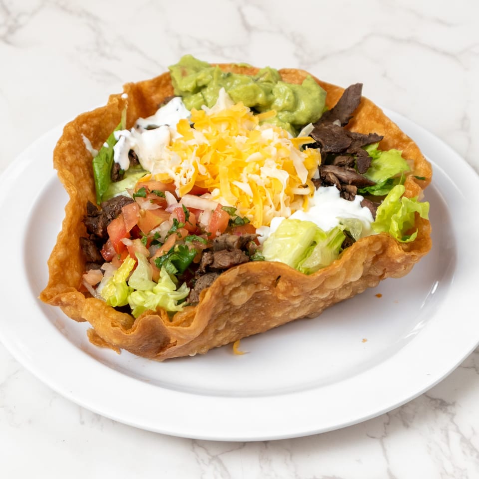Taco salad in a crispy fried tortilla bowl, filled with meat, lettuce, pico de gallo, cheese, sour cream, and guacamole.