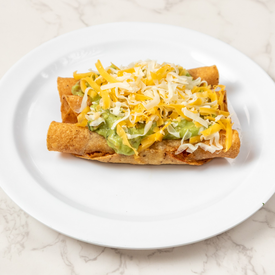 Three golden taquitos topped with guacamole and shredded cheese on a white plate.