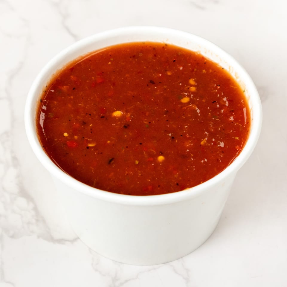A white cup filled with vibrant red salsa, featuring visible spices and small vegetable pieces.