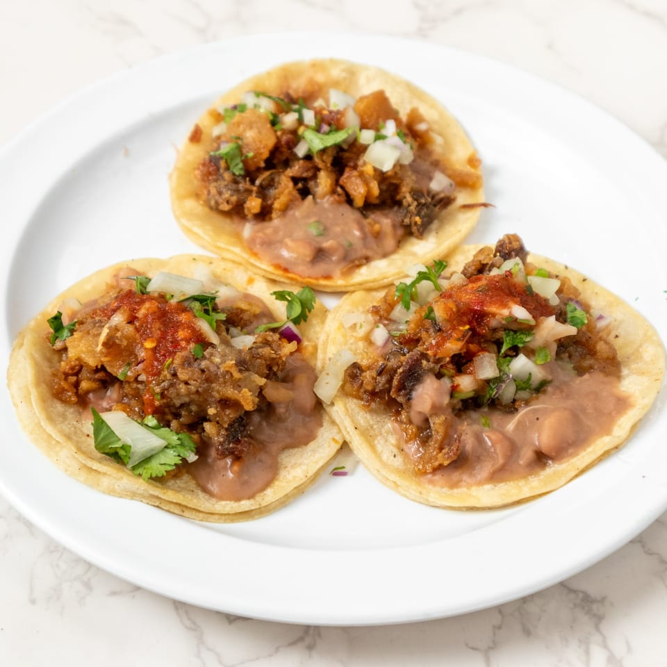 Three tacos with meat, refried beans, onions, cilantro, and red salsa on a white plate.