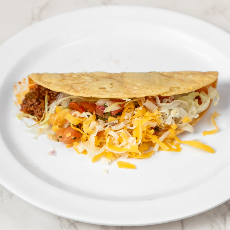 A single taco with ground beef, shredded cheese, lettuce, and diced tomatoes on a white plate.